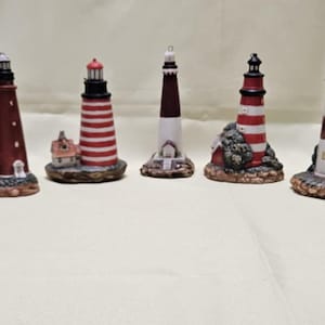 May include: A collection of eight miniature lighthouse figurines. The lighthouses vary in color, including red, white, and brown, with striped and solid designs. Each lighthouse has a detailed base, some with small houses or rocky textures.