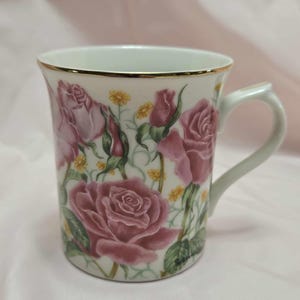 May include: White ceramic mug with a gold rim and a handle. The mug is decorated with pink roses, buds, and yellow flowers on a white background. The mug has a curved handle.
