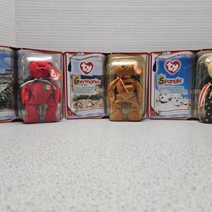 May include: Five Ty Beanie Baby plush toys in individual packaging. The toys are Osito, Germania, Spangle, and two others. The packaging features a red, white, and blue color scheme with the Ty logo and the name of the toy.