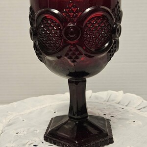 Op de afbeelding: Een robijnrode glazen kelk met een vierkante voet en een gedetailleerd, patroonontwerp. De kelk heeft een steel en een kom met een sierlijk, getextureerd patroon. Het glas heeft een vintage esthetiek.