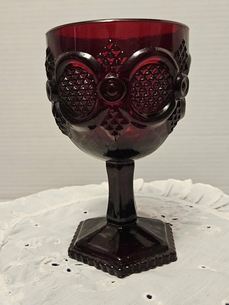Op de afbeelding: Een robijnrode glazen kelk met een vierkante voet en een gedetailleerd, patroonontwerp. De kelk heeft een steel en een kom met een sierlijk, getextureerd patroon. Het glas heeft een vintage esthetiek.