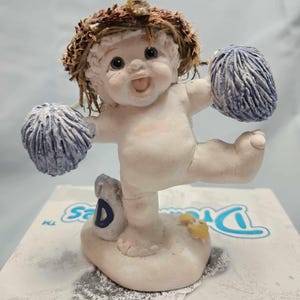 May include: A small, white figurine of a baby with brown hair wearing a blue and white pom-pom. The baby is holding a blue and white pom-pom in each hand and is standing on a base with the letter "D" on it.