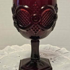 Op de afbeelding: Een robijnrode kelk met sierlijke details. Het glas heeft een ruitpatroon en decoratieve ovale vormen rond de kom. De kelk heeft een vierkante voet en een steel. Het glas is waarschijnlijk vintage of antiek.