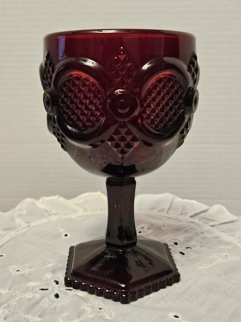 Op de afbeelding: Een robijnrode kelk met sierlijke details. Het glas heeft een ruitpatroon en decoratieve ovale vormen rond de kom. De kelk heeft een vierkante voet en een steel. Het glas is waarschijnlijk vintage of antiek.