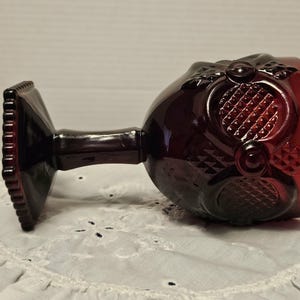Op de afbeelding: Een vintage robijnrood kelkglas met sierlijke details. Het glas heeft een vierkante voet, een slanke steel en een kom met een textuurpatroon. Het kelkglas rust op een wit, kantachtig oppervlak, dat de rijke kleur en het ontwerp benadrukt.