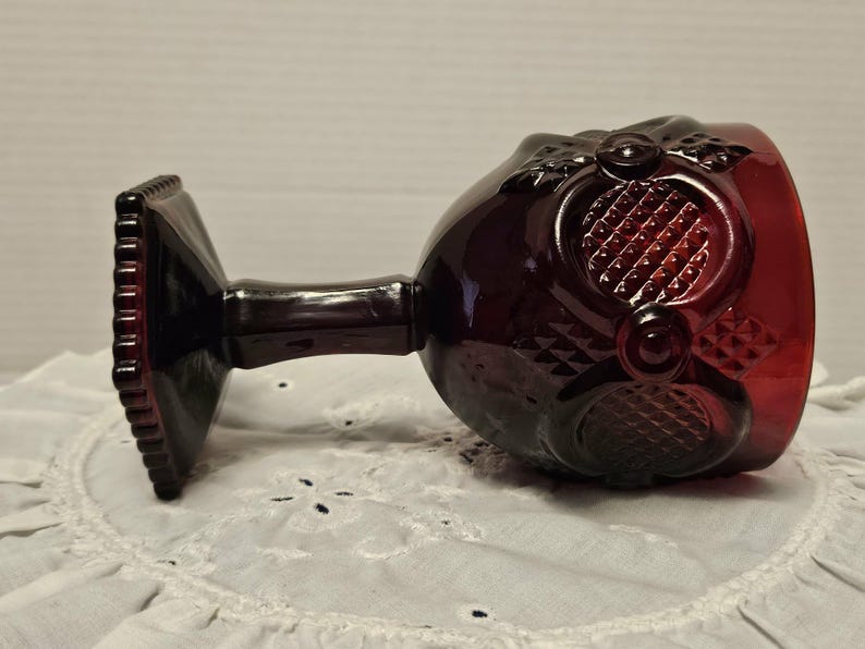 Op de afbeelding: Een vintage robijnrood kelkglas met sierlijke details. Het glas heeft een vierkante voet, een slanke steel en een kom met een textuurpatroon. Het kelkglas rust op een wit, kantachtig oppervlak, dat de rijke kleur en het ontwerp benadrukt.