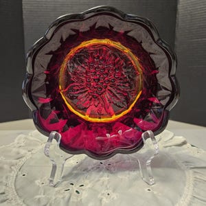 May include: A ruby red glass plate with a scalloped edge and a central floral design. The plate is displayed on a clear plastic stand, showcasing its intricate details and vibrant color. The plate's design is reminiscent of vintage glassware.