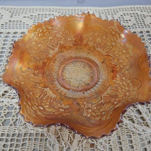 May include: A round marigold carnival glass bowl with a scalloped edge. The interior features a central textured honeycomb pattern surrounded by a relief design of blooming flowers and foliage with an iridescent, metallic orange finish.