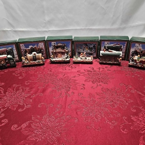 May include: A collection of six miniature Christmas-themed display boxes. Each box features a detailed diorama with figurines and miniature structures, set against a snowy backdrop. The boxes have green frames and are arranged on a red floral tablecloth.