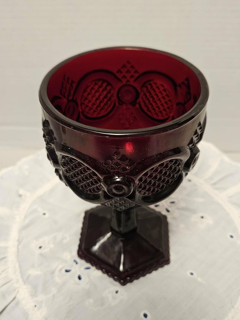 Op de afbeelding: Een vintage robijnrode kelk met ingewikkelde reli&euml;fpatronen. De kelk heeft een gedetailleerd ontwerp rond de kom en een zeshoekige basis. Het glas heeft een rijke, donkerrode kleur en de steel is ook rood.