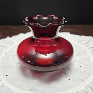 May include: A small, round, ruby-red glass vase with a ruffled rim. The vase has a bulbous body and a short neck. The rim is dark red, and the vase sits on a white, embroidered surface. The vase is likely for decorative purposes.