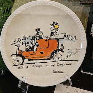May include: A white ceramic plate with a cartoon illustration of a vintage orange car carrying passengers. The plate has the text "walking through merrie Englande." and is signed "Norman Rockwell."
