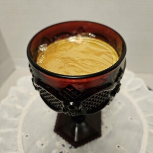 Op de afbeelding: Een robijnrode glazen beker met een donkere voet en een decoratief patroon rond de kom. De beker is gevuld met een lichtgele vloeistof. De beker staat op een wit, geborduurd oppervlak.
