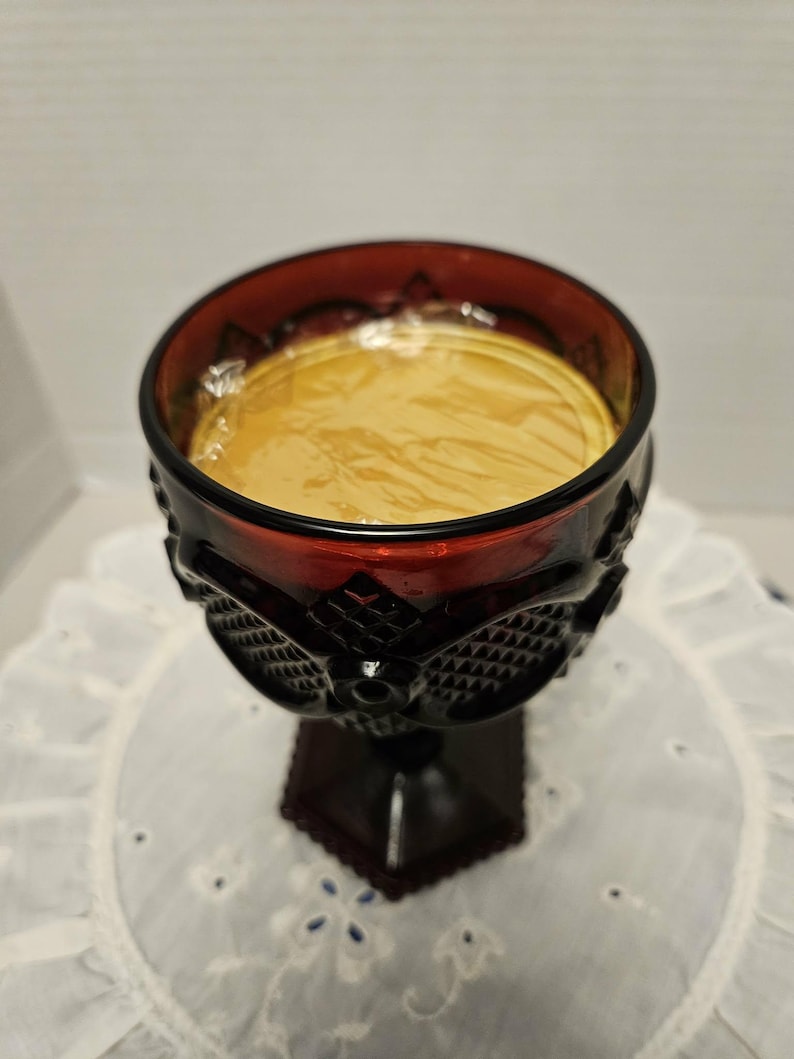 Op de afbeelding: Een robijnrode glazen beker met een donkere voet en een decoratief patroon rond de kom. De beker is gevuld met een lichtgele vloeistof. De beker staat op een wit, geborduurd oppervlak.