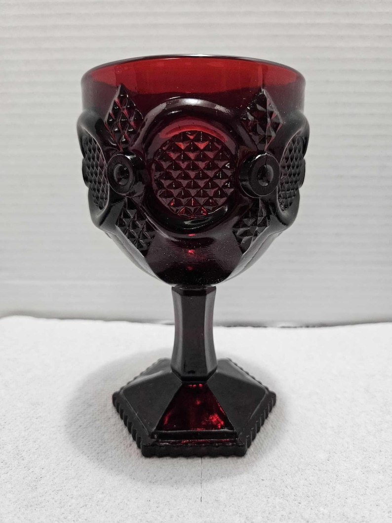 Op de afbeelding: Een robijnrode kelk met ingewikkelde reli&euml;fpatronen. Het glas heeft diamant- en cirkelvormige ontwerpen rond de kom, een steel en een geometrische basis. Het vintage glaswerk wordt waarschijnlijk gebruikt voor het serveren van wijn of andere dranken.