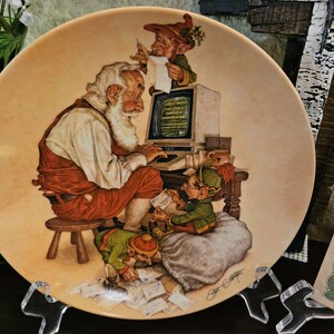 May include: A decorative plate featuring Santa Claus seated at a computer, surrounded by elves. The plate has a beige background with a detailed illustration. The text on the computer screen reads "GOOD NEWS AND GENTLE AMERICA".