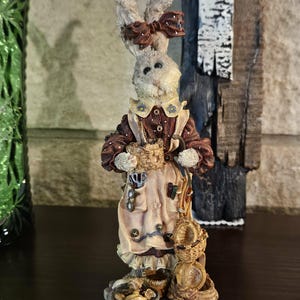 May include: A detailed figurine of a rabbit wearing a dress and apron, holding a basket. The rabbit has a bow on its head and is surrounded by small baskets and bowls. The figurine is painted in shades of brown, beige, and cream, with intricate details.