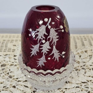 May include: A deep red glass votive holder with a textured surface and a white etched holly leaf and berry pattern. The red glass piece sits inside a clear, faceted glass base with a scalloped rim and a diamond-cut pattern on the exterior.