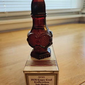 May include: A vintage Avon 1876 Cape Cod Collection salt shaker. The dark red glass bottle has an ornate design and a pointed stopper. The box reads "CHARISMA COLOGNE" and "1.5 FL.OZ."