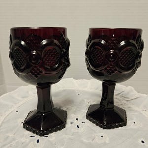 May include: Two ruby red vintage wine glasses. Each glass features a decorative pattern around the bowl and a faceted base. The stem and base are a dark, rich red. The glasses are displayed on a white, textured surface.
