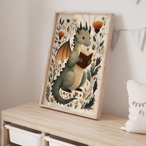 May include: A framed art print featuring a whimsical illustration of a green dragon reading a book, surrounded by flowers and foliage. The artwork has a light wood frame and is displayed on a wooden shelf. The overall theme is fantasy and children's decor.