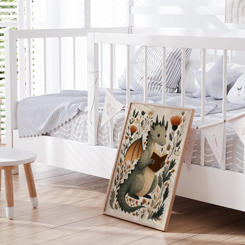May include: A framed print featuring a whimsical illustration of a green dragon reading a book, surrounded by flowers and foliage. The artwork has a light brown wooden frame. The print is leaning against a white crib with a gray and white patterned blanket.