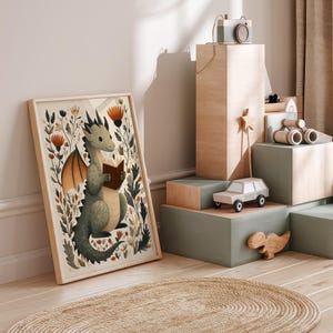 May include: A framed print featuring a whimsical illustration of a green dragon reading a book amidst floral elements. The artwork has a light wood frame and is displayed near wooden toy blocks and a toy car. The color palette includes greens, browns, and oranges.