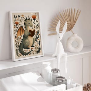 May include: A framed art print features a whimsical illustration of a green dragon reading a book, surrounded by flowers. A white bunny figurine sits nearby, along with a vase and decorative dried palm. The artwork is in a white frame.