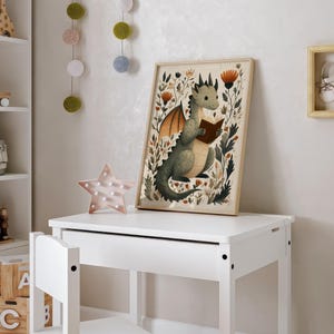 May include: A framed print featuring a whimsical illustration of a green dragon reading a book, surrounded by floral elements. The artwork has a light wooden frame and is displayed on a white desk. A pink star-shaped light and decorative hanging balls are also visible.