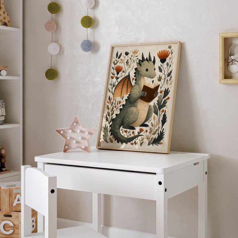May include: A framed print featuring a whimsical illustration of a green dragon reading a book, surrounded by floral elements. The artwork has a light wooden frame and is displayed on a white desk. A pink star-shaped light and decorative hanging balls are also visible.