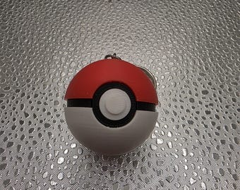 May include: A spherical keyring designed as a red and white ball with a black horizontal band across the centre. A circular black and white button is positioned in the middle of the sphere. A small metal ring is attached to the top of the object.