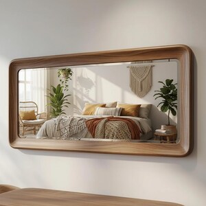 Puede incluir: Un gran espejo de pared rectangular con un marco de bordes redondeados y acabado de veta de madera natural. La superficie reflectante muestra el interior de un dormitorio con una cama, plantas en macetas, una silla y un tapiz de pared.