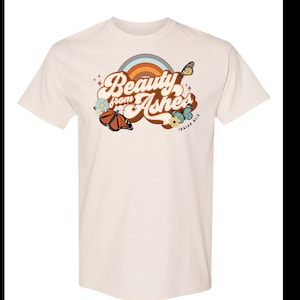 May include: A beige t-shirt with a retro-style graphic design featuring the text "Beauty from Ashes" with a rainbow, butterflies, and flowers. The text "ISAIAH 61:3" is also included on the shirt.