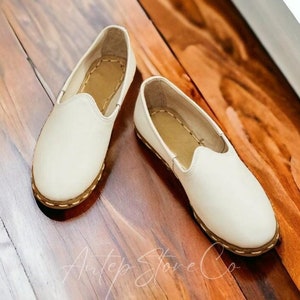 Pode incluir: Um par de sapatos brancos de couro sem cadarços com uma borda dourada ao redor das bordas. Os sapatos estão em um piso de madeira.
