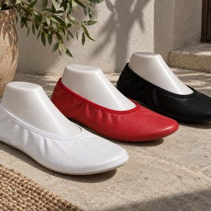 Può includere: Tre ballerine slip-on esposte su forme per piedi bianche. Le scarpe sono presentate nei colori bianco, rosso e nero, ciascuna caratterizzata da punta arrotondata, scollo basso e un design semplice e privo di decorazioni con suola piatta.