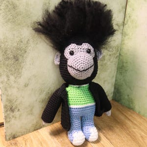 Puede incluir: Una muñeca de mono de ganchillo hecha a mano con un afro negro, cara gris y una expresión sonriente. La muñeca lleva una camisa verde, vaqueros azules, una chaqueta negra y zapatos blancos. El juguete está sobre una superficie de madera.