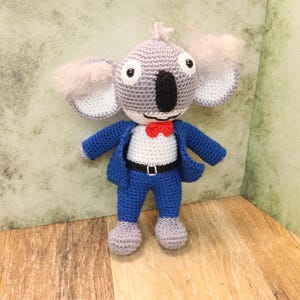 Puede incluir: Koala de ganchillo con un traje azul, camisa blanca y pajarita roja. El koala tiene pelaje gris, orejas grandes y una nariz negra. El juguete está sobre una superficie de madera.