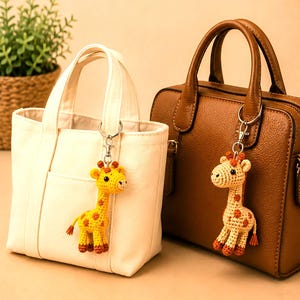 Può includere: Due portachiavi a forma di giraffa all'uncinetto attaccati a delle borse. Uno è giallo brillante con macchie arancioni, appeso a una borsa tote in tela bianca. L'altro è beige chiaro con macchie marroni, appeso a una borsa in pelle marrone.