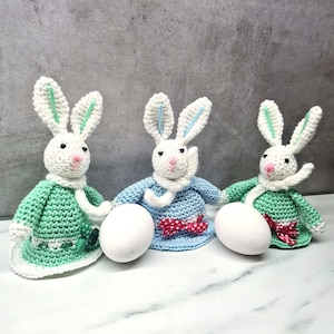 May include: Three crocheted bunny rabbit figurines holding white eggs. The bunnies are wearing green, blue, and green dresses with white collars and red bows. The bunnies are sitting on a white marble surface.