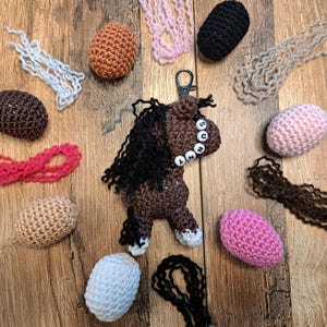 May include: A brown and black crocheted horse keychain with the name "SUNNY" spelled out in white beads. The image also includes various crocheted horse parts in different colors, such as pink, brown, and white, on a wooden surface.