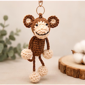 May include: A crocheted monkey keychain featuring a brown body and head with cream-colored accents on the face, ears, hands, and feet. The monkey has small black eyes and a stitched black mouth, attached to a gold-toned metal ring and clasp.