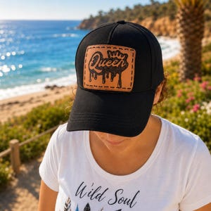 May include: A black baseball cap featuring a rectangular tan leather patch on the front. The patch is stitched with black thread and displays the word Queen in a stylized script font with a crown icon above it and a dripping paint effect below.