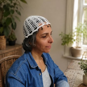 Op de afbeelding: Een witte gehaakte schedelkap met een opengewerkt rasterpatroon en een rij kleine, ronde metalen bedeltjes die langs de onderrand hangen. Het hoofddeksel is gemaakt van fijn wit garen met een delicaat, herhalend geometrisch ontwerp.