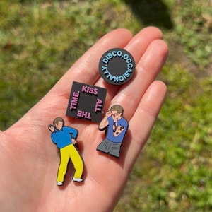 May include: Four colourful shoe charms are displayed. One is a black circle with the text "DISCO, OCCASIONALLY" in blue. Another is a black square with "KISS ALL THE TIME" in pink. The other two charms are cartoon figures.
