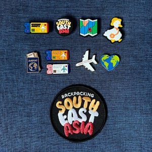 May include: A selection of travel-themed pins and patches. Includes a passport, airplane tickets, a map, a duck with a travel bag, a heart-shaped globe, an airplane, and a patch that says "Backpacking South East Asia."