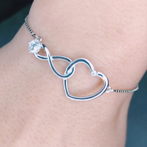 May include: A silver bracelet featuring a double heart design with a single rhinestone at the centre. The bracelet is made from a delicate chain.