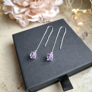 Könnte beinhalten: Ein Paar silberne Ohrhänger mit einem linearen Design. Jeder Ohrring hat einen kleinen, hellvioletten, würfelförmigen Kristall. Die Ohrringe werden auf einer schwarzen Schachtel präsentiert, mit einem weichen, verschwommenen Hintergrund.