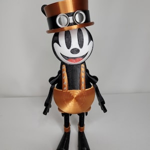 Op de afbeelding: Een zwart-wit cartoonfiguur met een brede, tanden grijns, die een bruine hoed met een bril en een bruin en zwart pak draagt. Het figuur staat op een wit oppervlak.