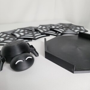 Könnte beinhalten: Schwarze Spinnenfigur mit weißen Augen und ein schwarzes Spinnennetz-Untersetzer-Set. Die Spinnennetz-Untersetzer sind übereinander gestapelt.