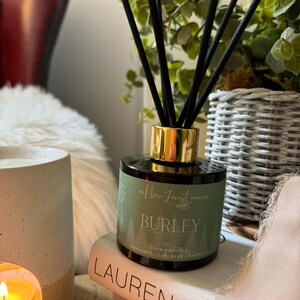 May include: A reed diffuser with black reeds and a gold-coloured collar. The green label reads "Burley" and lists fragrance notes. The diffuser sits on a book with the title "Lauren Conrad". A lit candle and a decorative plant are also visible.